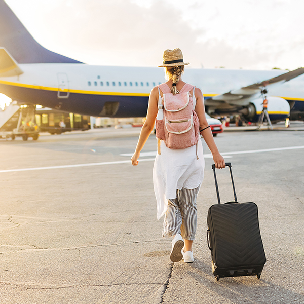 Frau mit Strohhut, Rucksack und Rollkoffer geht auf ein Flugzeug zu